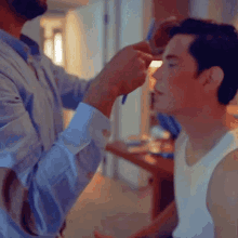 a man is applying makeup to another man 's face while wearing a white tank top .