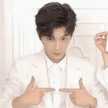 a man in a white suit and tie is pointing at himself with his hands .