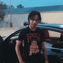 a young man wearing a black t-shirt with a picture of a baby on it is standing next to a car .