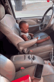 a baby is sitting in the driver 's seat of a car with a phone in the center console