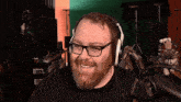a man with a beard wearing headphones and glasses smiles in front of a green screen
