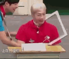 a man in a red shirt is sitting at a desk while another man stands behind him