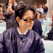 a man wearing sunglasses and a blue jacket is sitting in a crowd of people .