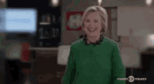 hillary clinton is wearing a green sweater and smiling while standing in a room .