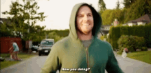 a man in a green hoodie is standing on a street and smiling while asking how you doing .