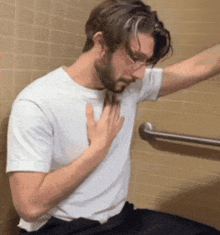 a man with a beard and glasses is sitting in a bathroom with his hands on his chest