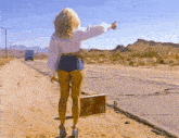 a woman walking down a dirt road with a suitcase giving a thumbs up