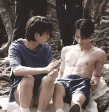a shirtless man sits next to another shirtless man on a blanket