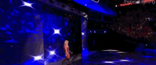 a wrestler is walking on a stage in a dark room with a blue light behind him .