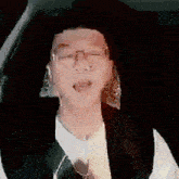 a man wearing glasses and a vest is sitting in a car with his hands in his hair .