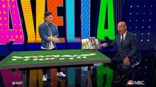 two men are sitting at a table with cards on it in front of a nbc sign