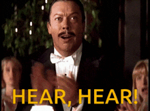 a man in a tuxedo applauds with the words hear hear in yellow letters