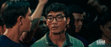 a man wearing glasses is talking to another man with kahar written on the bottom right