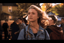 a woman in a denim jacket stands in a crowd of people with her hair blowing in the wind