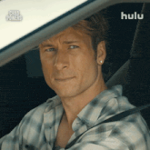 a man in a plaid shirt is sitting in the driver 's seat of a car with a hulu logo in the background