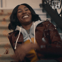 a woman in a brown leather jacket is smiling in front of a sign that says be