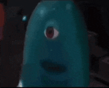 a blue monster with a red eye is looking at the camera in a dark room .