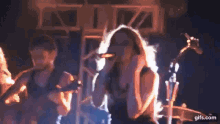 a woman is singing into a microphone on a stage while a man plays a guitar in the background .