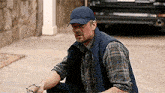 a man wearing a hat and vest is kneeling on the sidewalk