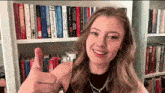 a woman giving a thumbs up in front of a bookshelf full of books including one called a rising man