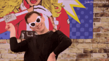 a man wearing sunglasses and a black shirt is standing in front of a brick wall with a picture of a woman on it
