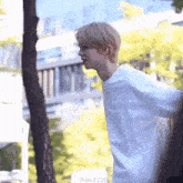 a young man in a white shirt is standing next to a tree and talking to someone .