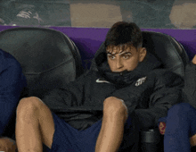 a man is sitting in a purple chair with his legs crossed and a nike jacket on .