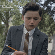 a boy in a suit and tie is writing with a pencil in front of a son of a witch logo