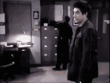 a man in a black jacket is standing in a room with a man in a black jacket standing in the background .