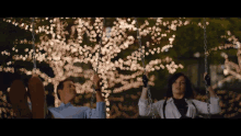 a man and a woman are sitting on swings in front of a tree with lights on it