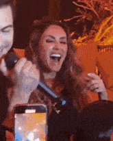 a woman is singing into a microphone while a man takes a picture of her with a cell phone .