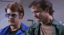 two men wearing sunglasses and blue jackets are standing next to each other in a room .