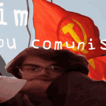 a man wearing glasses holds a piece of paper in front of a red flag with the words " im ou communis " written on it