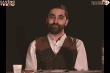 a man with a beard sits at a table with a saving throw wildcards logo behind him