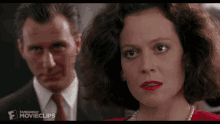 a man in a suit and tie stands next to a woman in a red dress and pearls