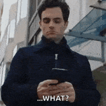 a man in a blue jacket is looking at his cell phone and says `` what '' .