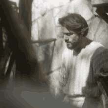 a man with a beard wearing a white shirt is standing in front of a stone wall