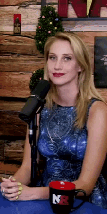 a woman in a blue dress sits in front of a microphone next to a red mug that says n8