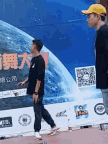 a man wearing a yellow hat is standing next to another man in front of a large sign with chinese characters on it