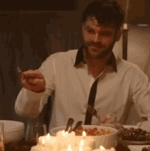a man in a white shirt and tie is sitting at a table with candles and food .