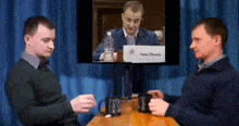 two men are sitting at a table talking to each other in front of a television screen .