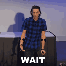 a man in a blue plaid shirt is holding a microphone and the word wait is above him