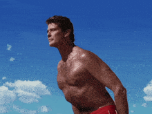 a shirtless man in red swim trunks stands on a beach