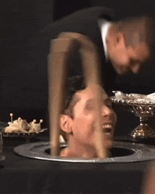 a man is sticking his head through a hole in a table while another man looks on .
