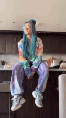 a girl with blue hair and glasses is sitting on a counter in a kitchen .