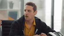 a man wearing a yellow shirt and a black jacket is sitting in front of a phone