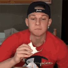 a man wearing a yankees hat and a red champion shirt is eating a sandwich