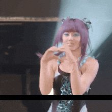 a woman with purple hair and blue eye shadow is making a heart shape with her hands