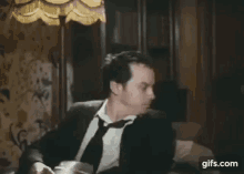 a man in a suit and tie is sitting on a couch with a lamp in the background .