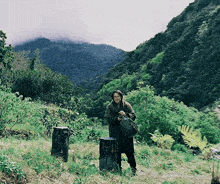 a woman standing in a field with trees and stump in the foreground and mountains in the background